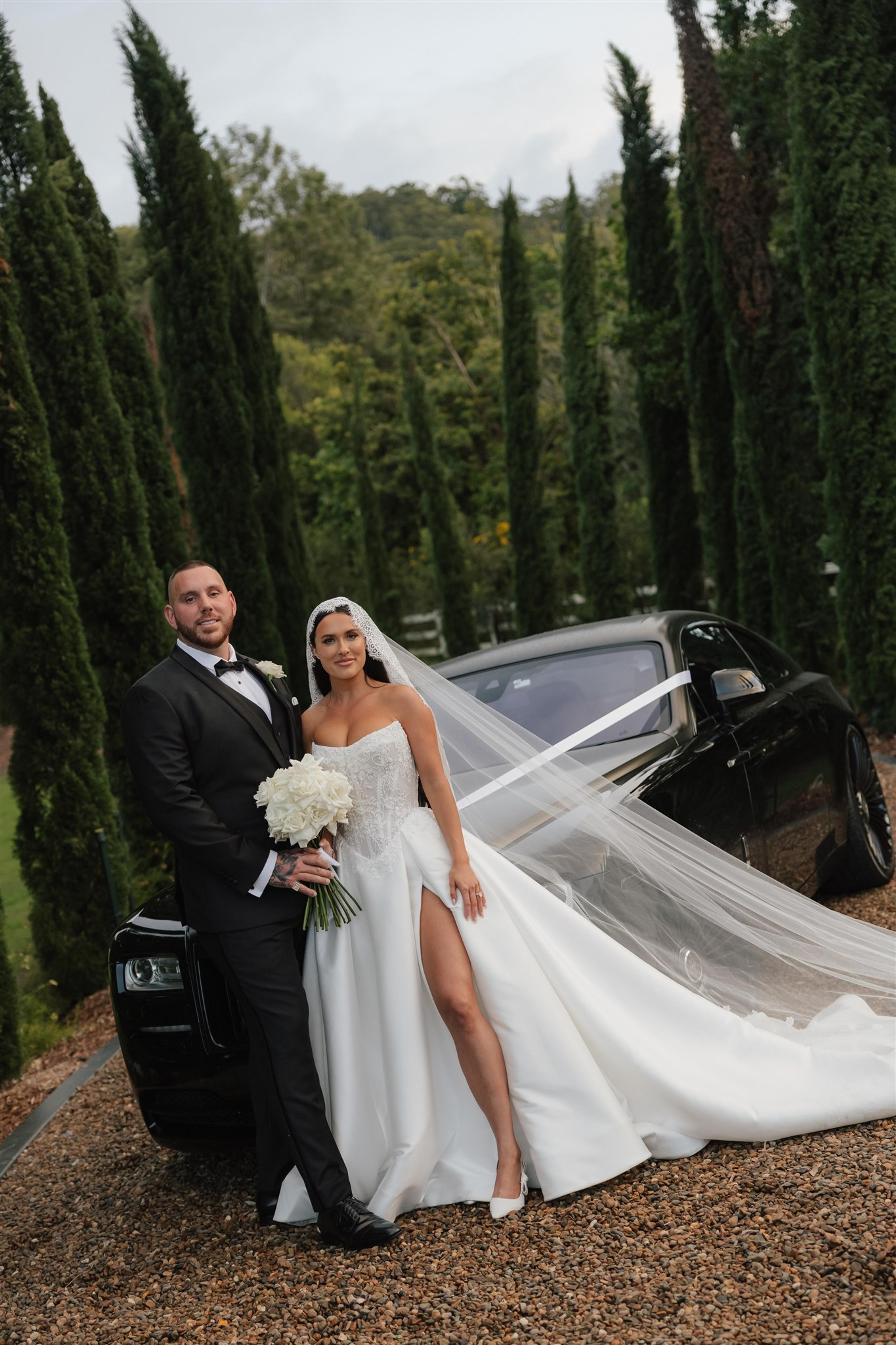 Rochelle and Jeff posing beside a black wedding car at The Valley Estate Gold Coast, with the bride holding a white rose bouquet.