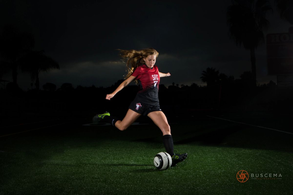 Young female soccer player in red and black uniform kicking ball on night field