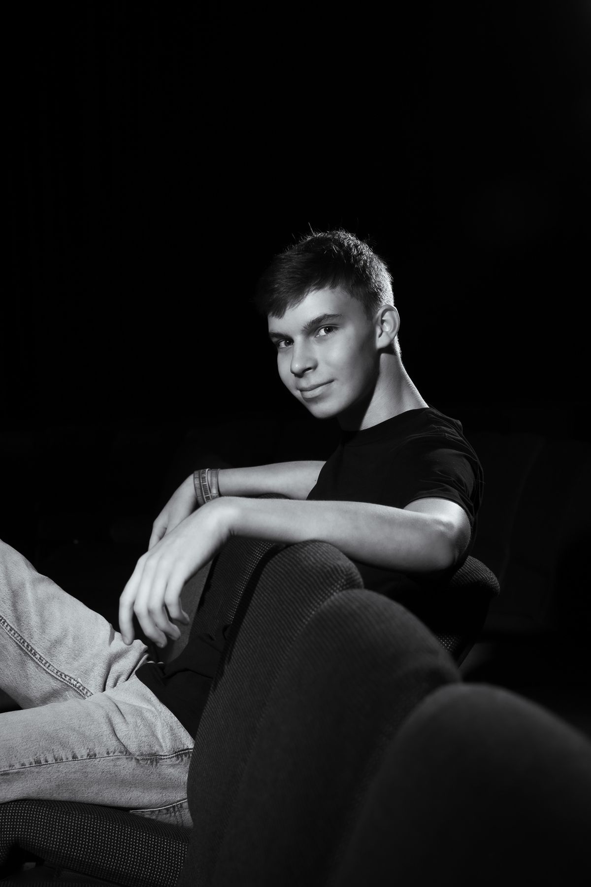 Black and white senior portrait of a young man in casual pose against dark background, looking at camera with slight smile.