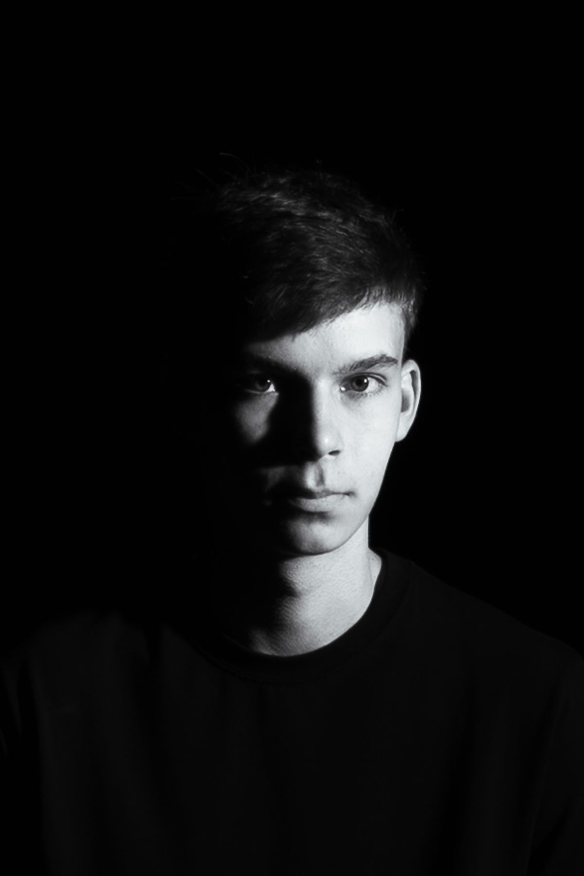 Black and white portrait photograph of a young man against a dark background, looking directly at the camera with a serious expression.