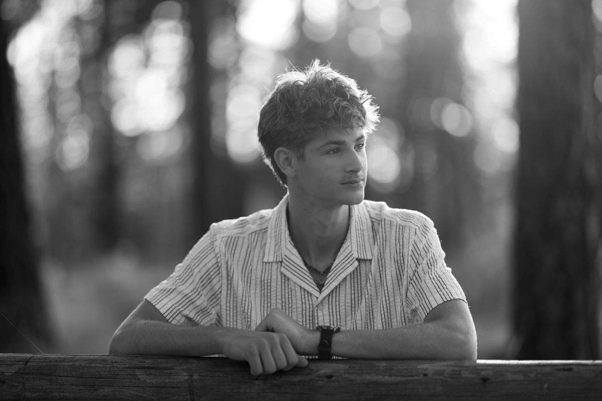 Black and white senior portrait of a Coeur d'Alene student leaning on a wooden rail among the trees