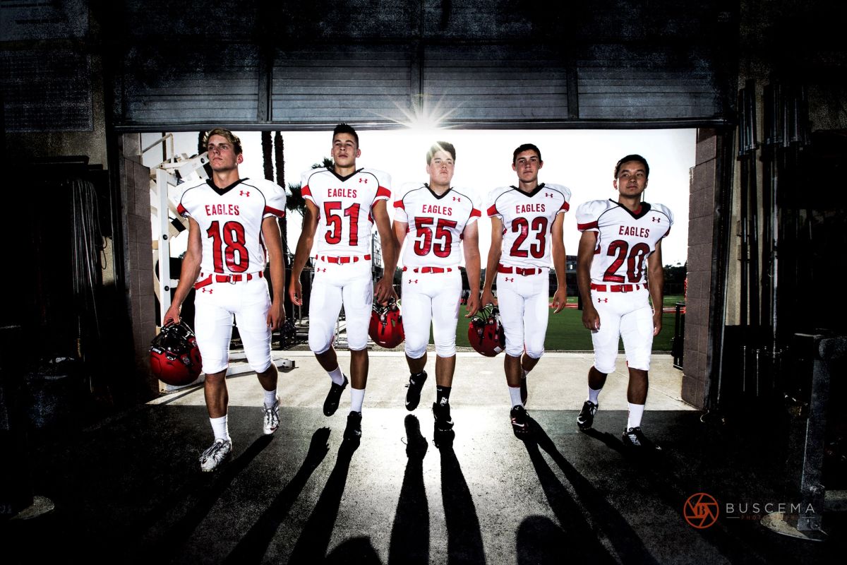 Five Eagles football players in red and white uniforms walk out of a tunnel toward the field, carrying helmets.