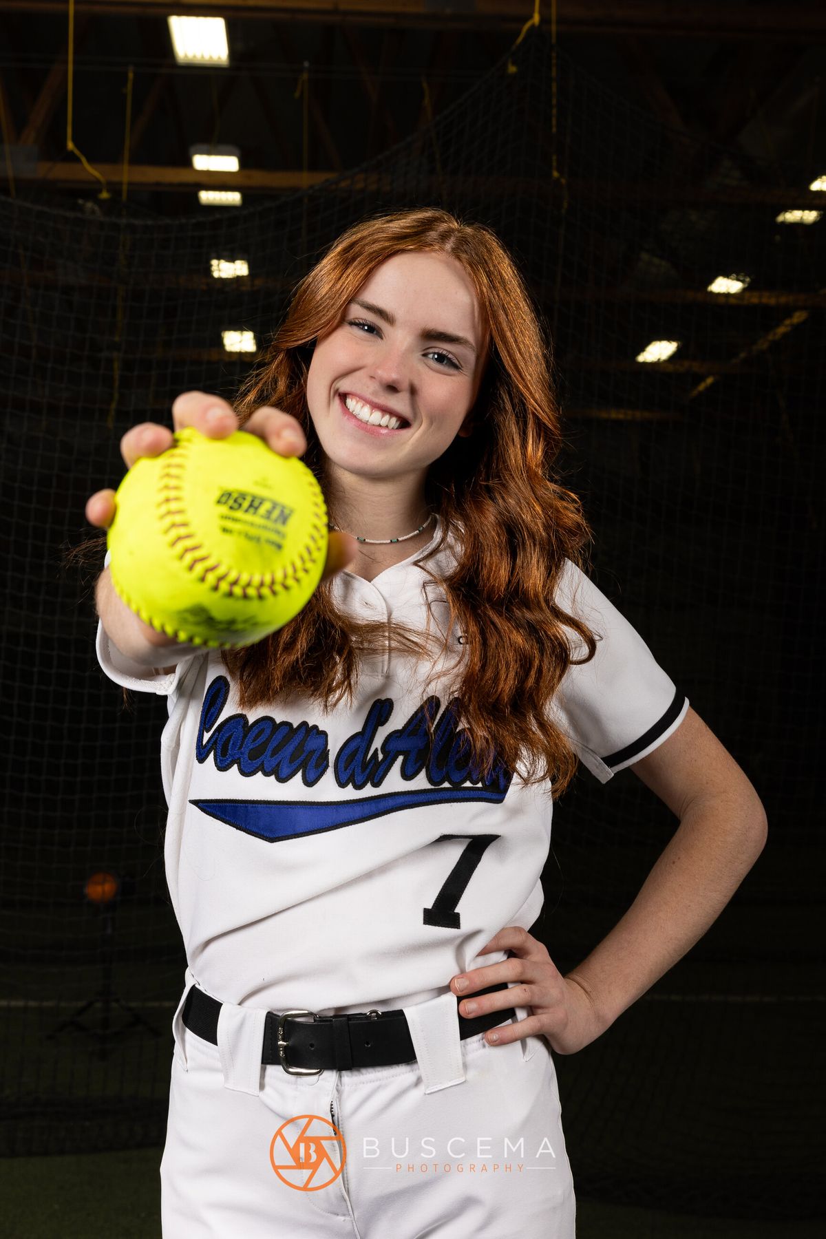 Female softball player wearing white uniform with blue "Wildcats" script and number 7, holding yellow softball, smiling at camera.