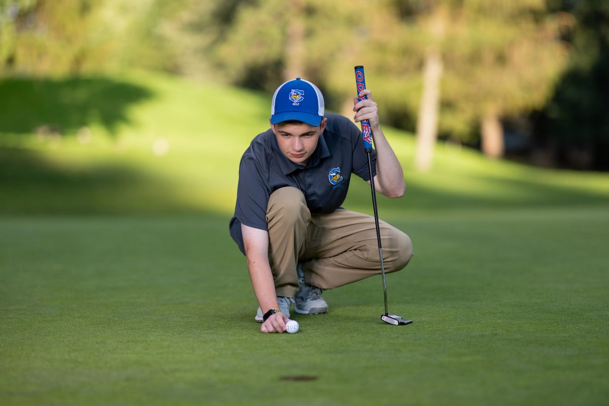 Young golfer in blue cap crouches on putting green, lining up putt with putter and golf ball.