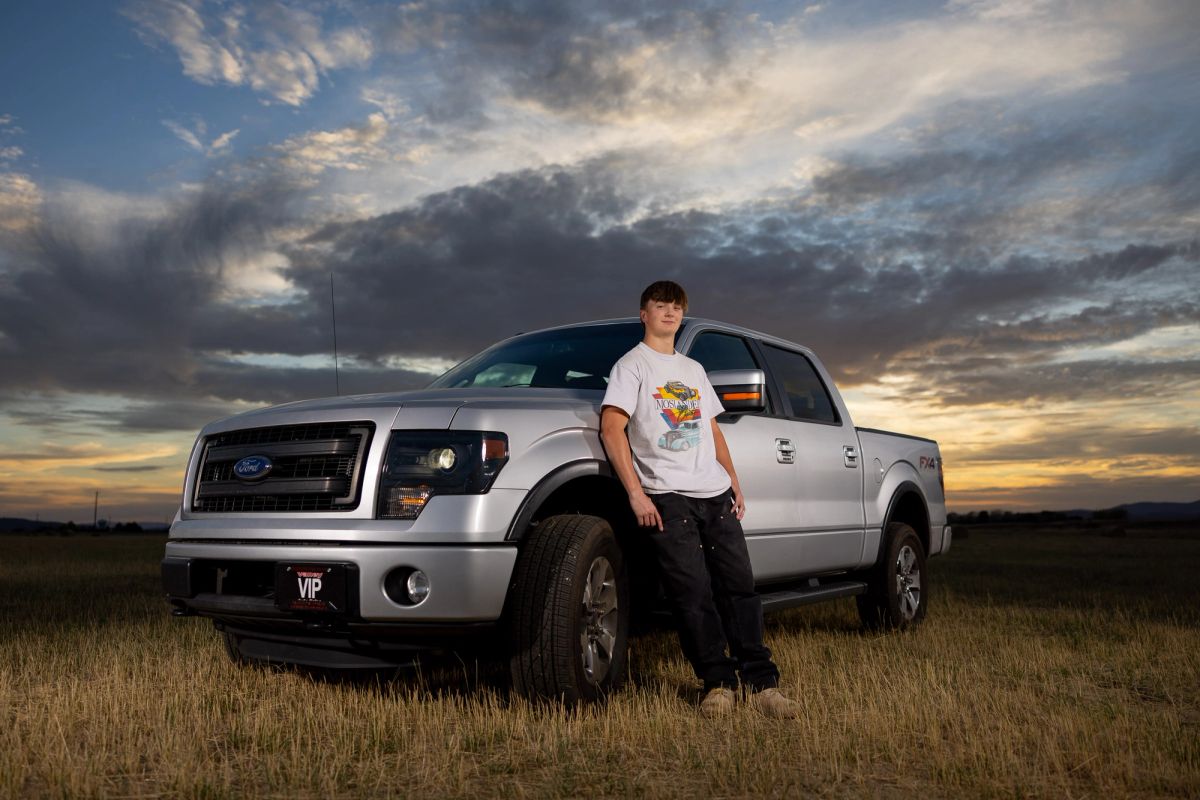 Mason Chapman poses leaning against a silver Ford F-150 pickup truck in a grassy field under a dramatic cloudy sky.