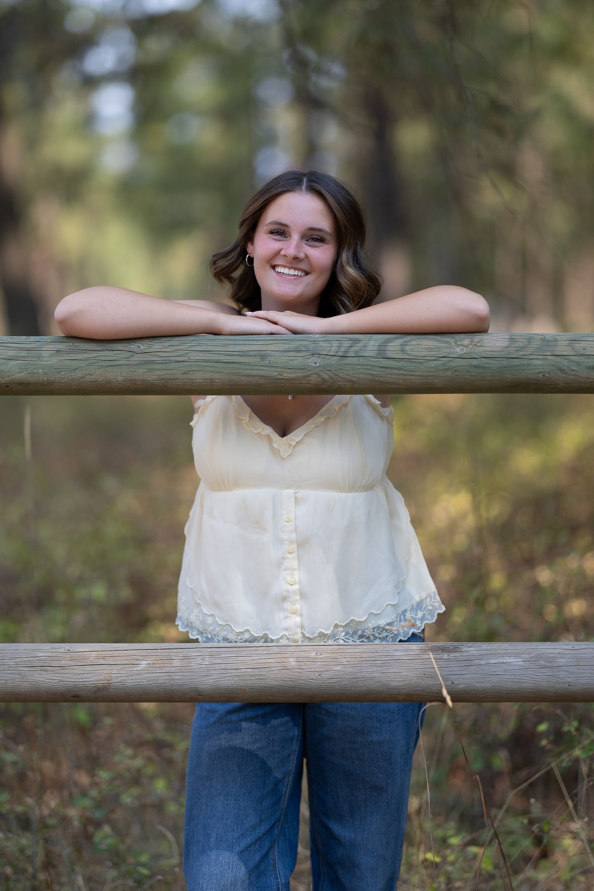 Smiling young woman leaning on wooden fence rail in forest setting, wearing cream top and jeans