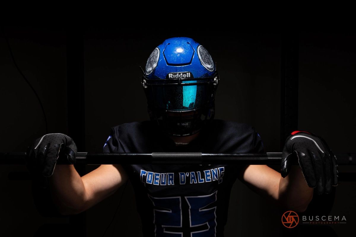 Football player in blue Riddell helmet holding barbell, wearing "Coeur d'Alene" jersey against black background.