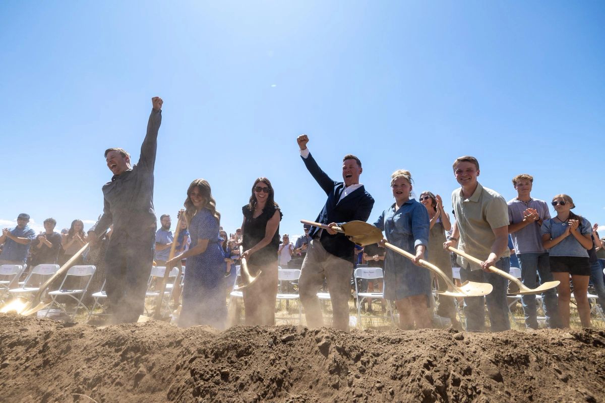 Group of people in formal attire holding gold shovels at a groundbreaking ceremony, standing in dirt with dust clouds and spectators in background.