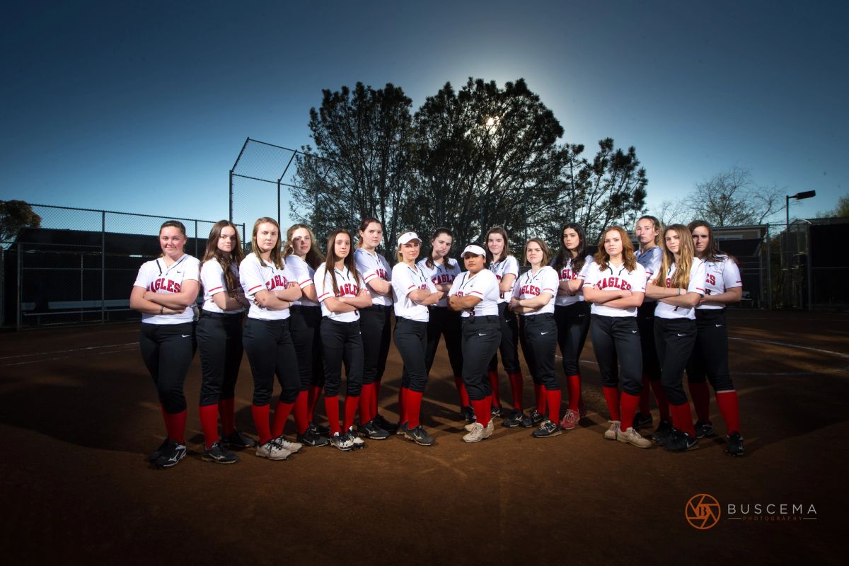 Eagles softball team posed in uniform on dirt field with baseball diamond and trees in background.