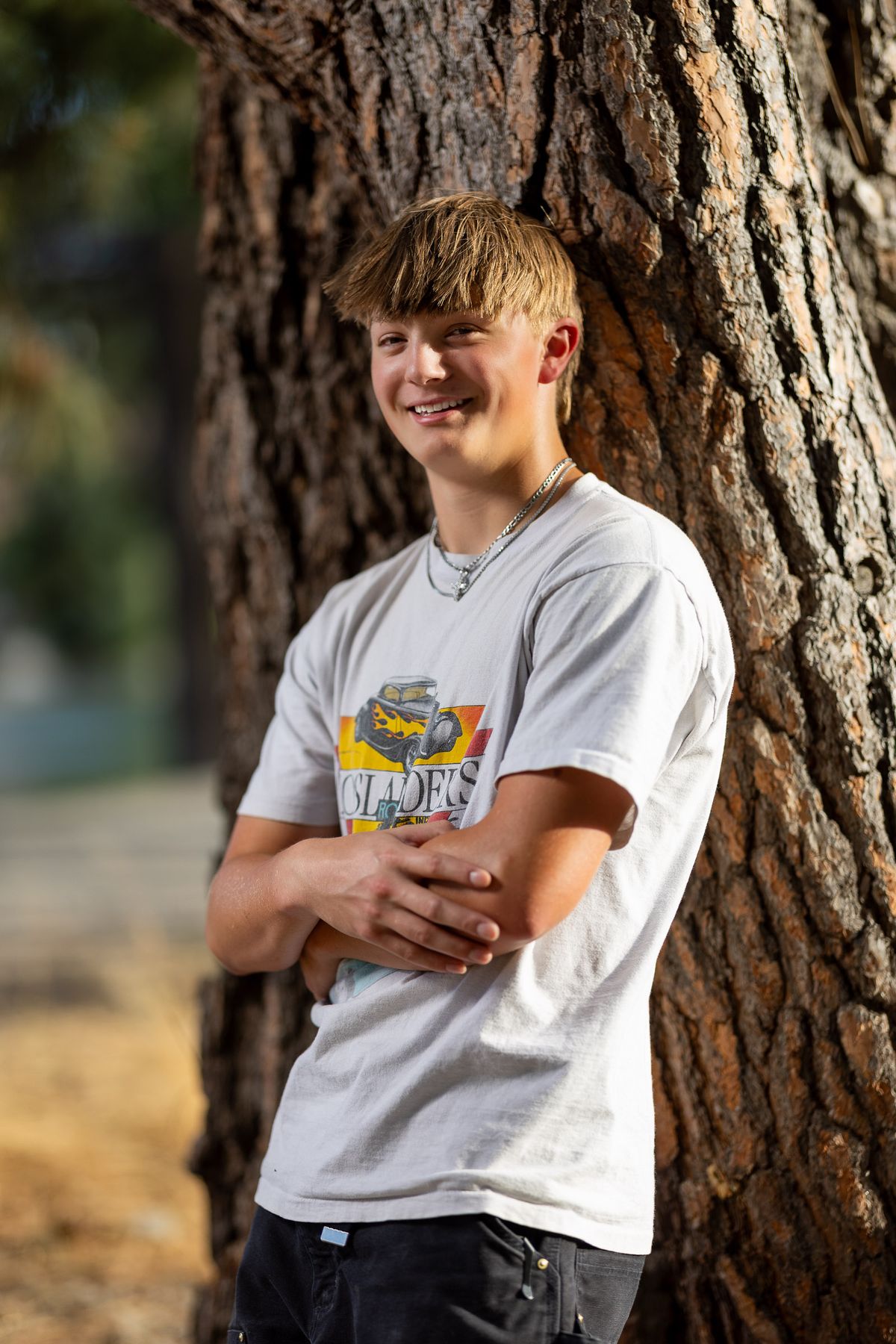A smiling teenage boy in a gray graphic t-shirt leans against a large tree trunk in outdoor natural lighting.