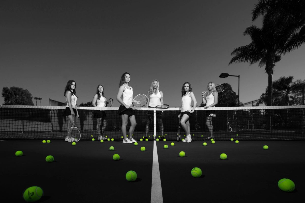 Six female tennis players stand at the net holding rackets with tennis balls scattered on the court in this black and white senior portrait.