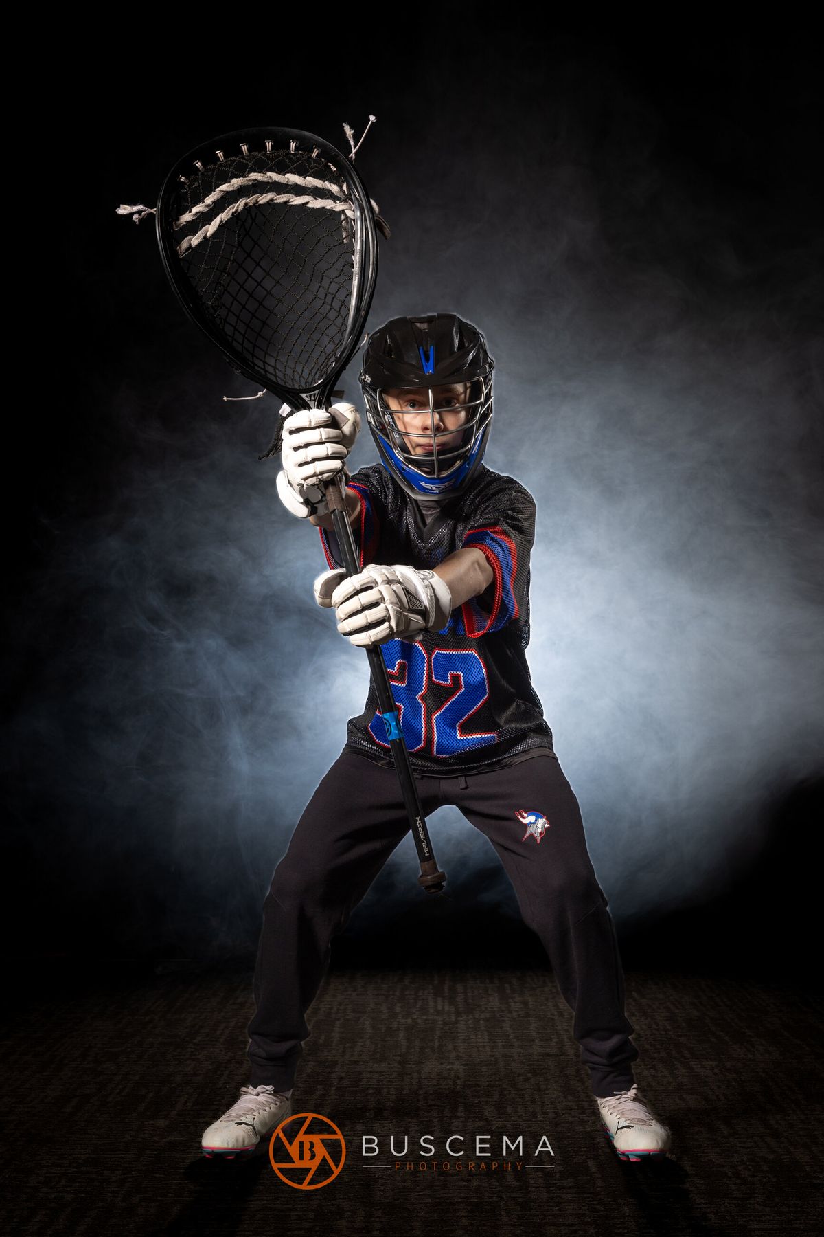Young lacrosse player wearing blue helmet and #32 jersey, holding stick in athletic portrait pose.