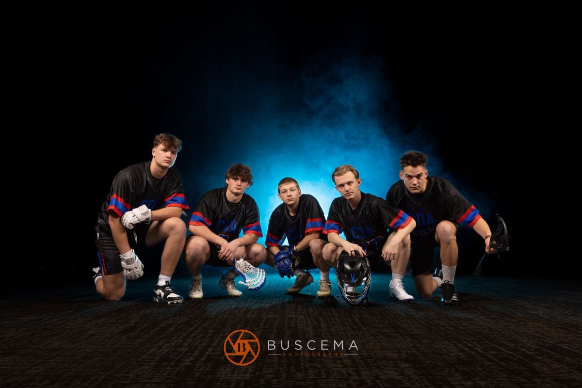 Five male lacrosse players in black and red uniforms crouching together against a blue-lit background, holding helmets and gloves.
