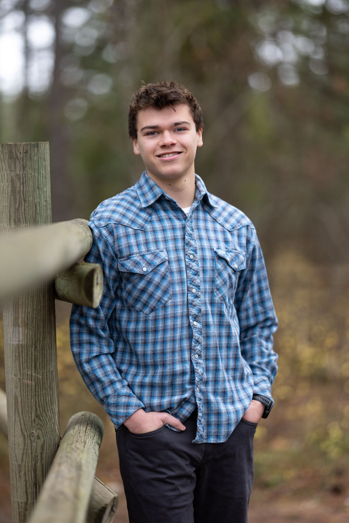 High school senior portrait of Ty Van Meeteren in blue plaid shirt leaning against wooden fence