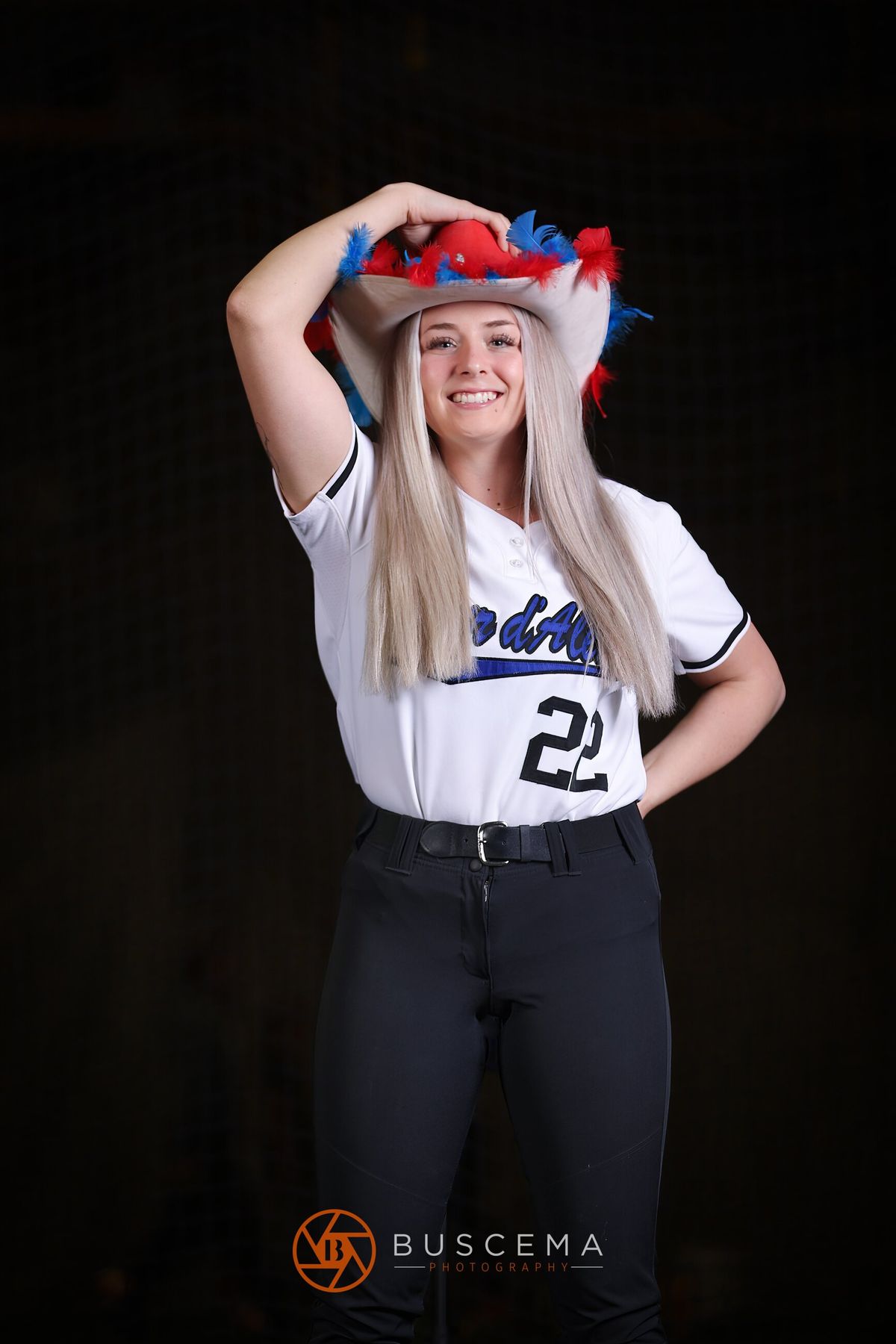 Smiling female athlete in white #2 jersey wearing patriotic feathered cowboy hat against black background.