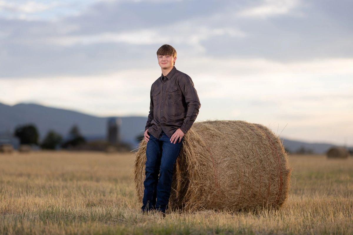 A young man in a dark shirt and jeans stands beside a large round hay bale in a harvested field with mountains visible in the distance.