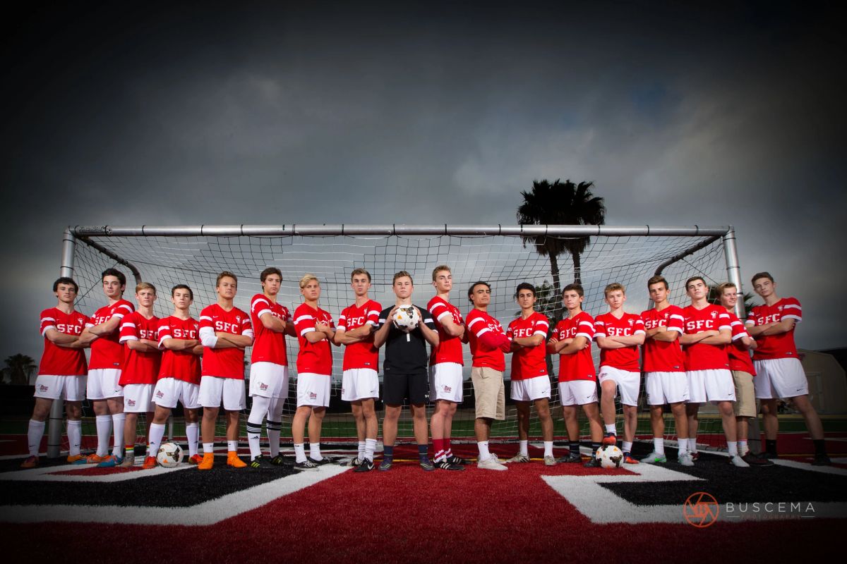 Soccer team in red and white uniforms lined up in front of goal net with palm tree and dramatic sky.