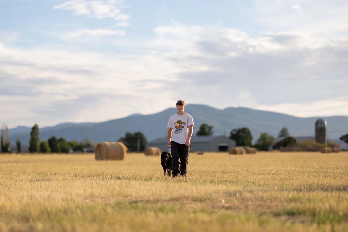Young man walking with black dog through harvested field with hay bales and mountains in background.
