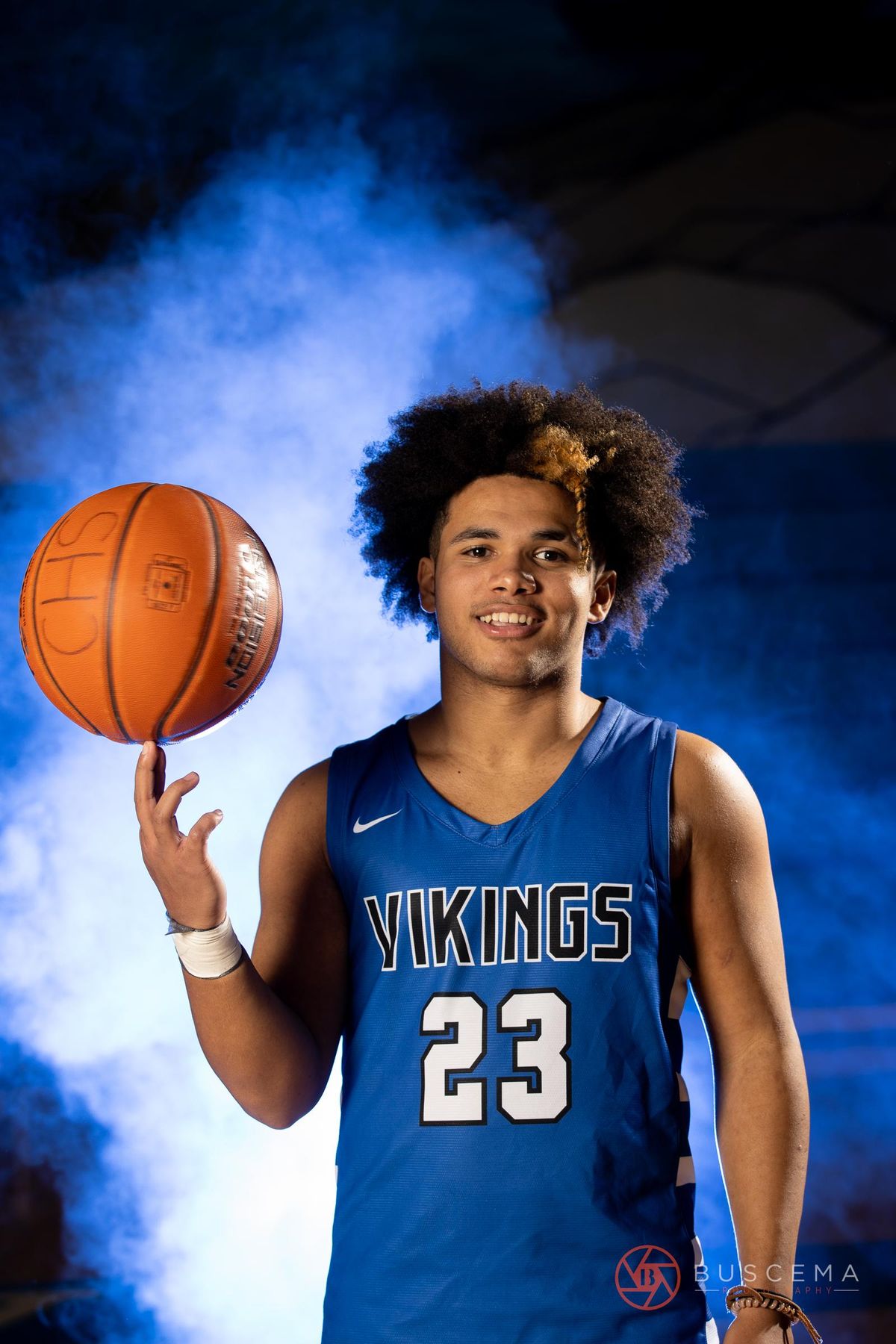 Young basketball player wearing Vikings #23 jersey spinning orange ball on finger against blue background.