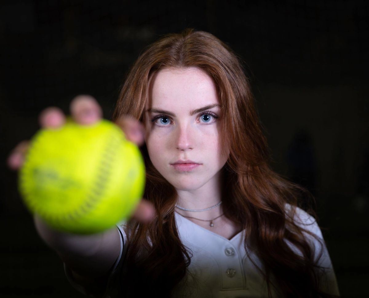 Young female athlete holding a bright yellow softball, looking directly at camera with blurred teammates in background.
