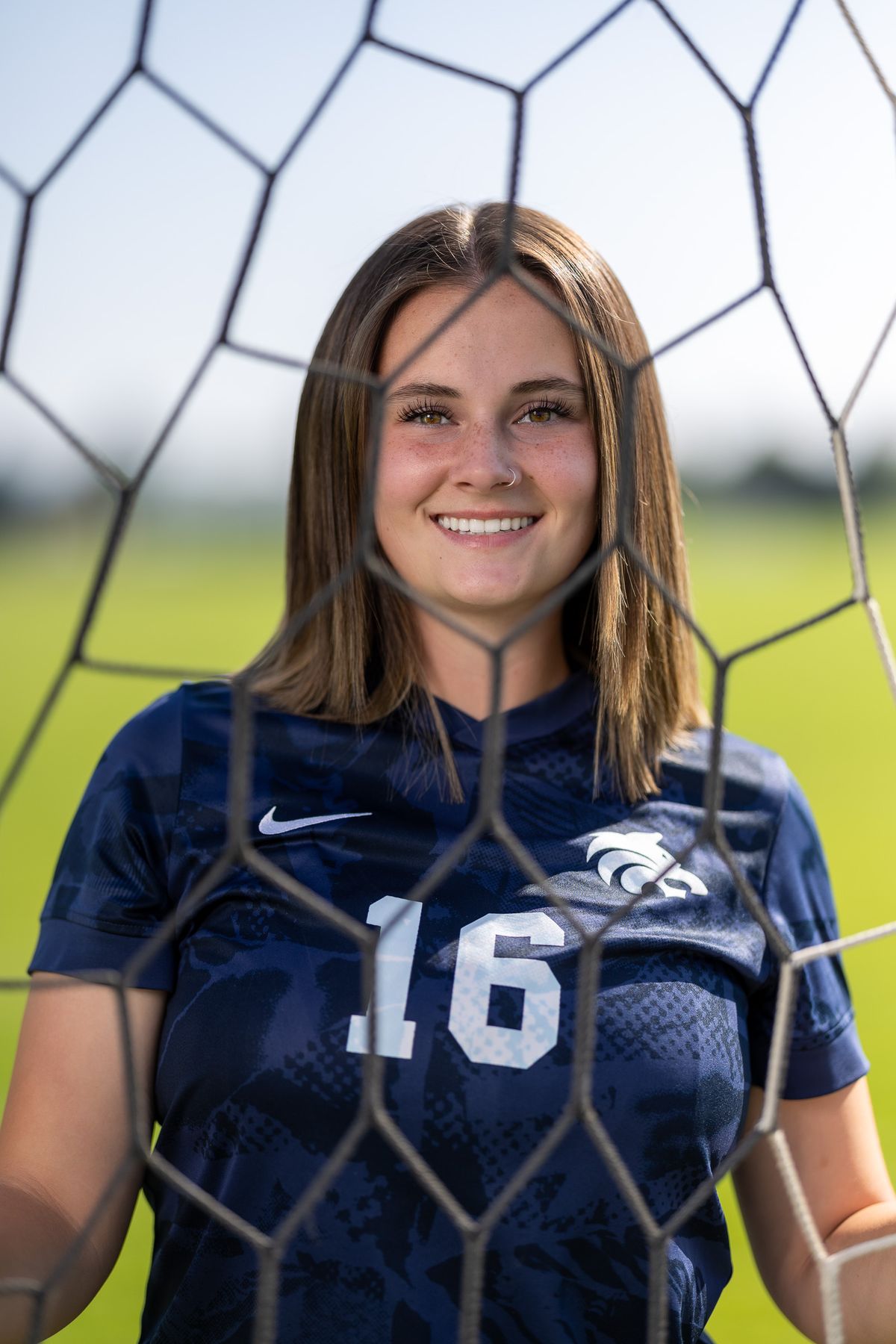 High school soccer player wearing navy jersey number 16 posed in front of goal net with field background.