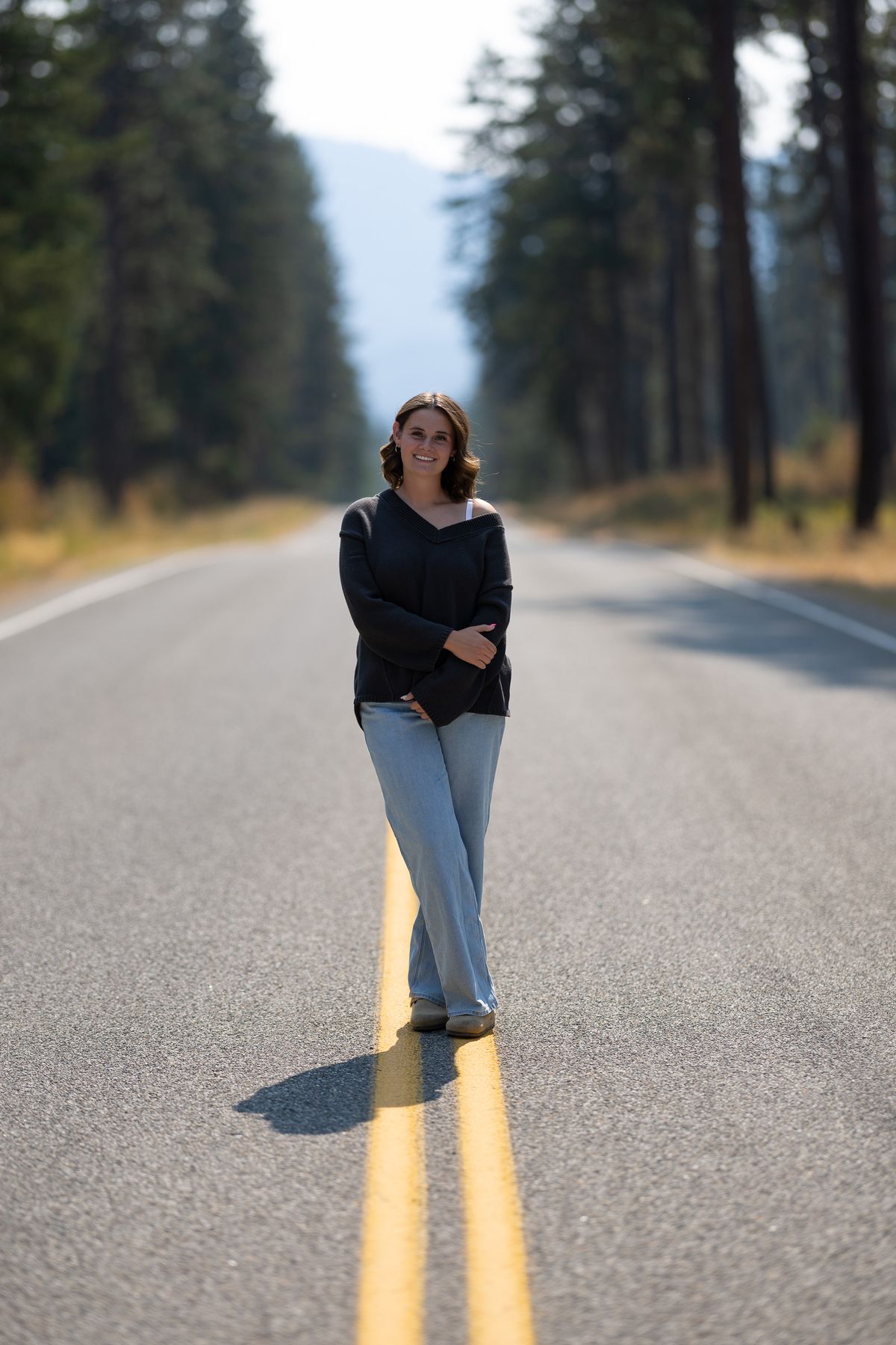 A young woman standing on a straight road lined with tall pine trees, smiling at the camera with mountains visible in the distance.