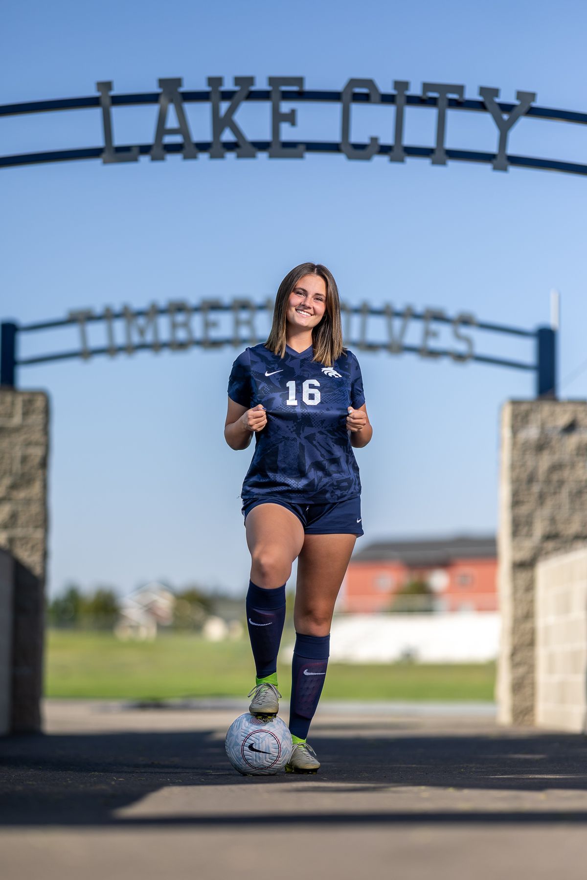Young female soccer player in navy uniform number 16 with ball under Lake City High School sign.