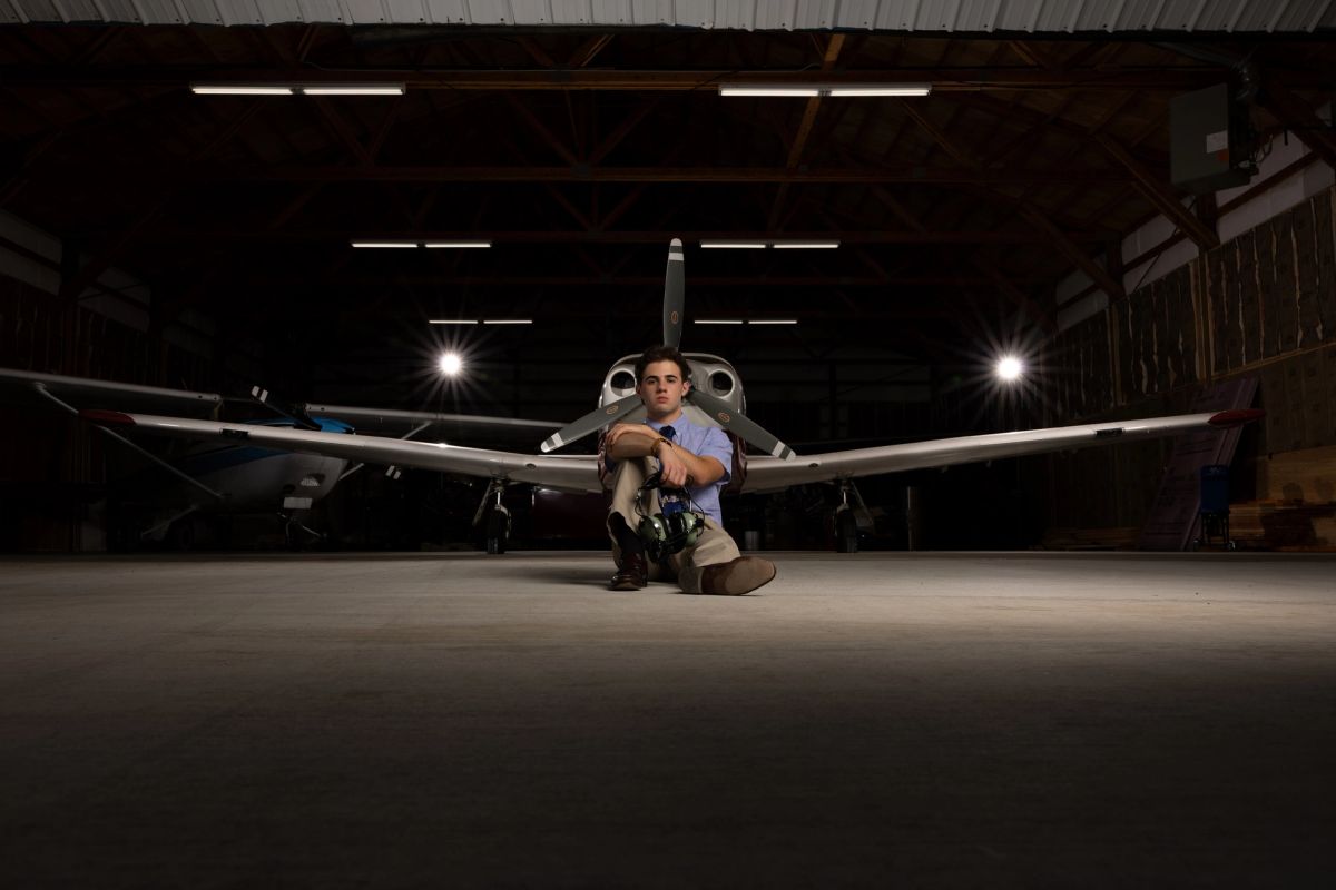 Coeur d'Alene high school senior kneeling in front of a small airplane in a lit hangar