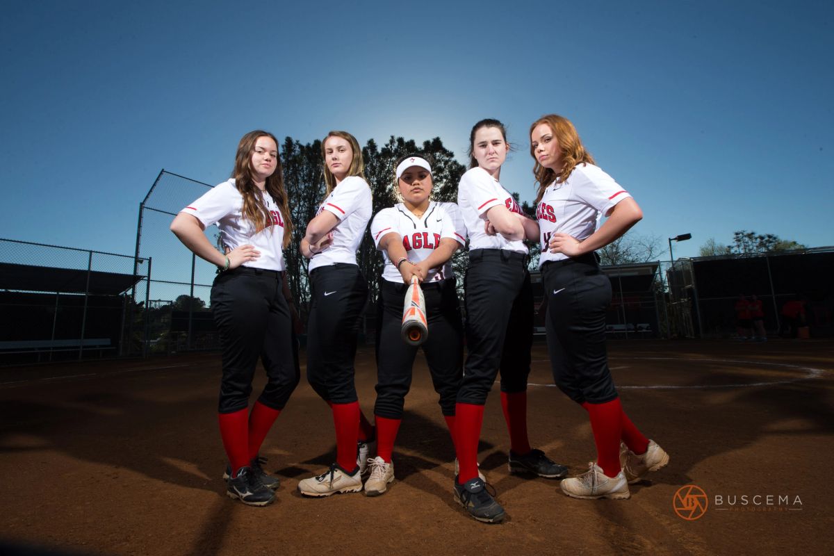 Five female softball players in white and red uniforms posing together on a dirt field under clear blue sky.