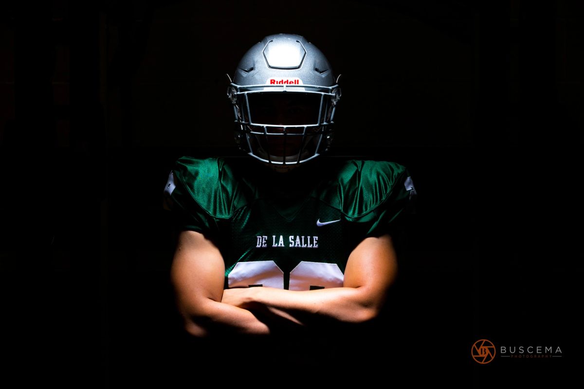 Football player in De La Salle green jersey and silver Riddell helmet holding football against black background.