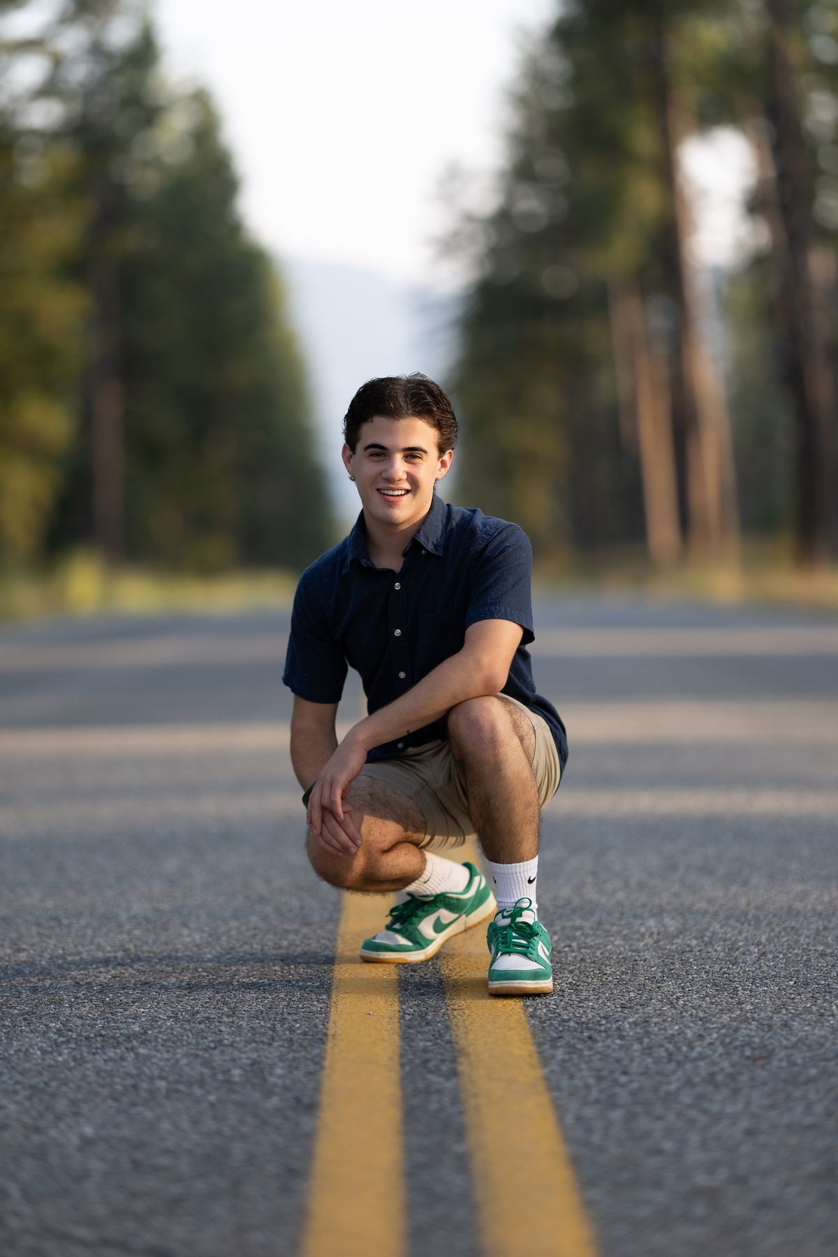 Coeur d'Alene high school senior kneeling on a road centerline with evergreens behind him