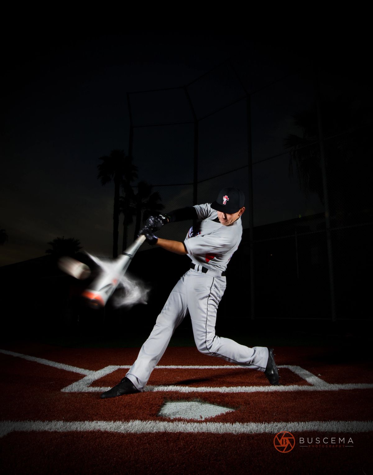 Baseball player in gray uniform mid-swing at night, with motion blur on bat and ball, against dark stadium background.