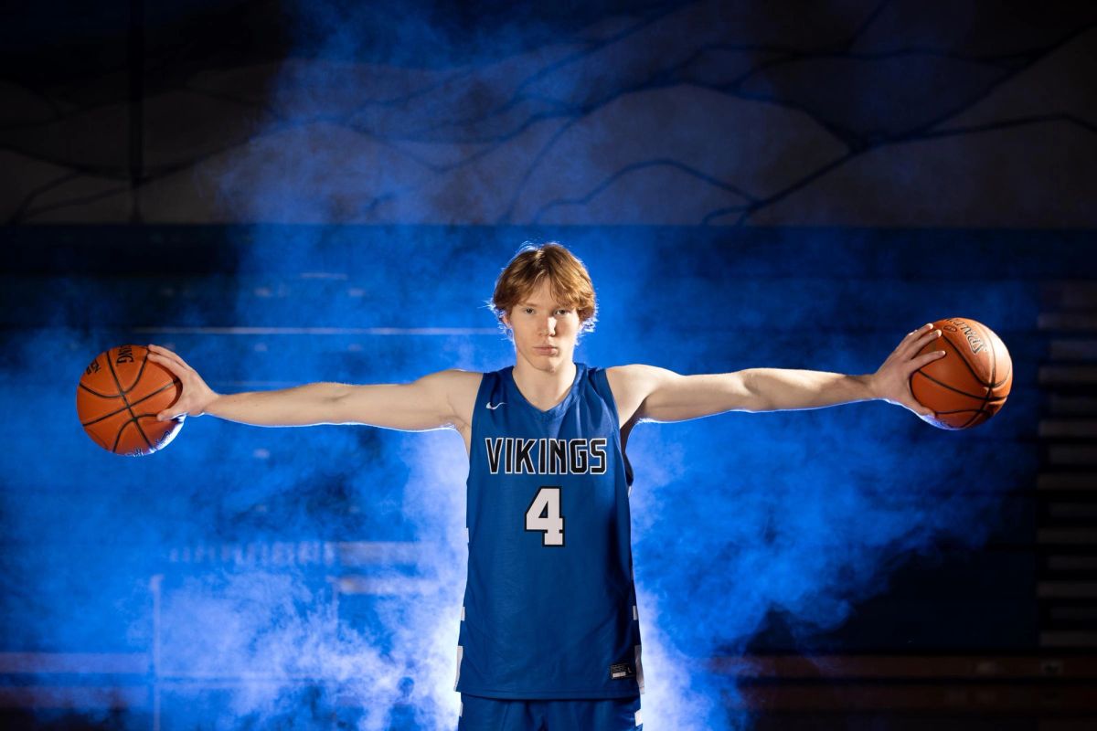 Young basketball player wearing Vikings #4 jersey holds basketballs with arms extended against blue backdrop.