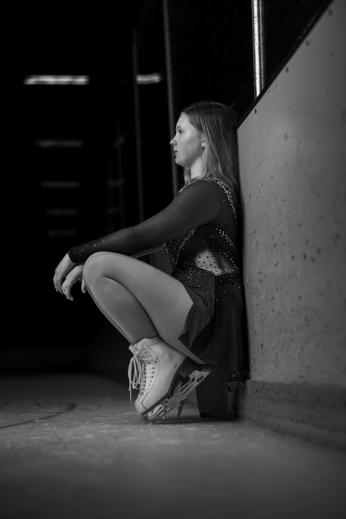 Black and white senior portrait of a figure skater crouched against a concrete wall in her skates