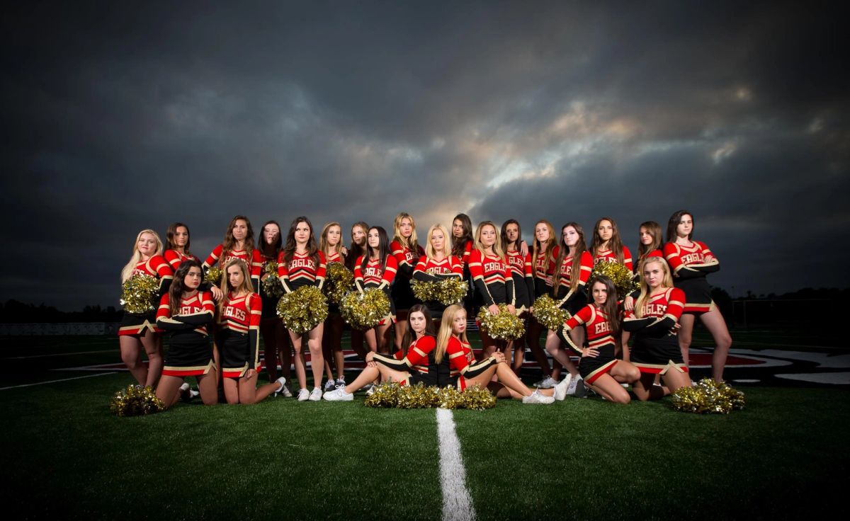 Coeur d'Alene High School cheerleaders in red and gold uniforms with pom-poms posed on football field under dramatic sky.