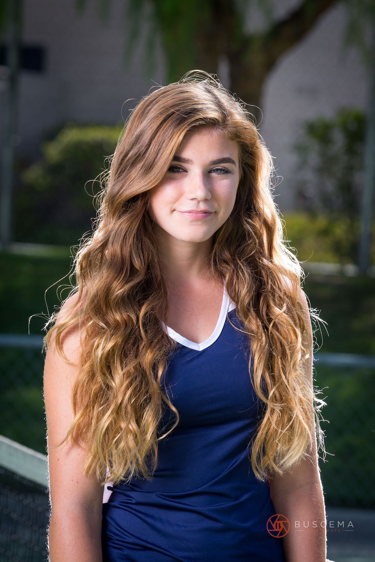 Young woman with long wavy auburn hair wearing a dark blue V-neck tank top, photographed outdoors with blurred greenery background.