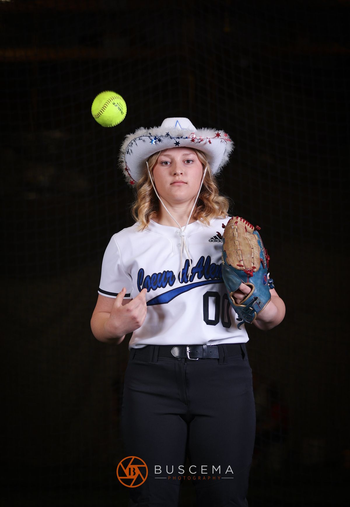 Young softball player in white uniform and patriotic cowboy hat holding glove, with yellow ball floating above head against dark background.