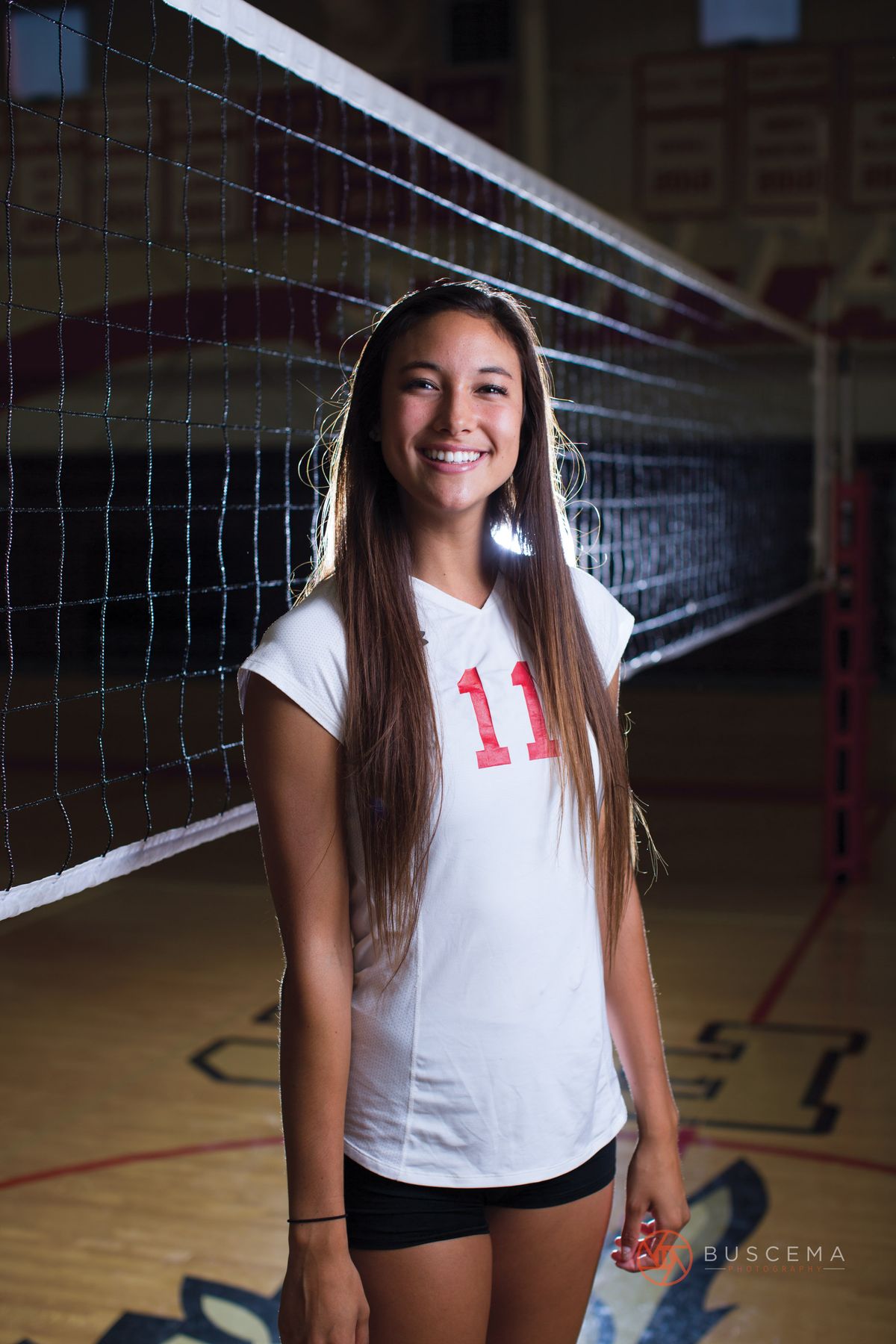 High school volleyball player wearing number 11 jersey posing by net in gymnasium.