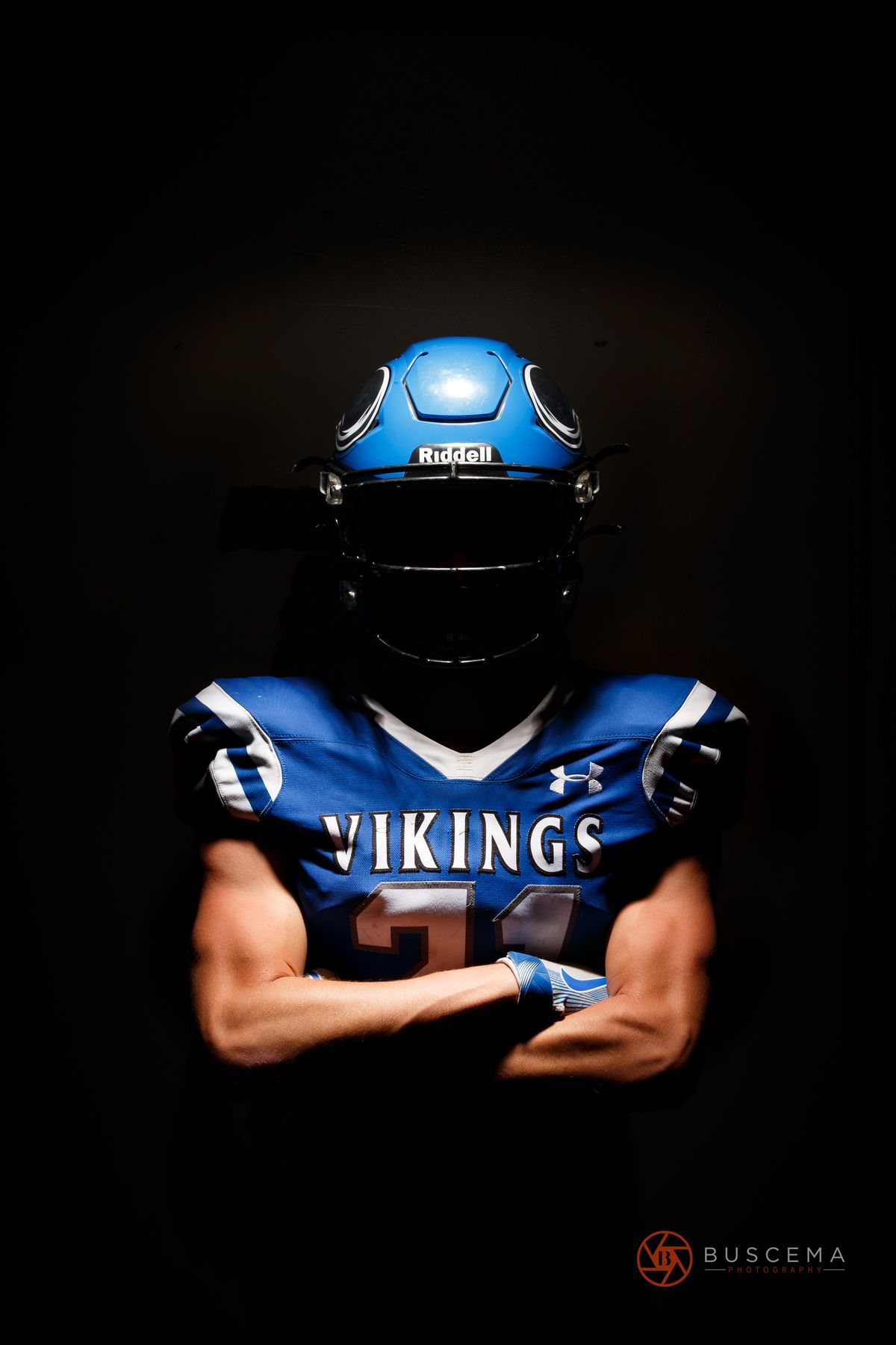 Football player wearing blue Vikings jersey and Riddell helmet with arms crossed against black background.