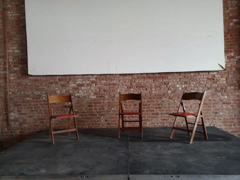 Three wooden folding chairs sitting under a projection screen on a small stage