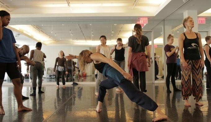 One woman in a dance pose as multiple other women dance in a well lit mirrored room