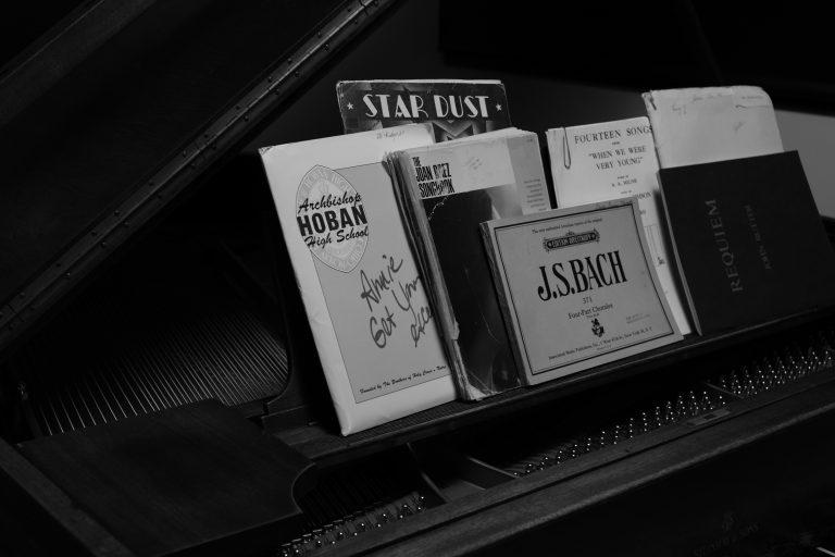 Black and white image of a piano with numerous books of classic sheet music on top. Sheet music from composer J.S. Bach, the play "Annie Get Your Gun", and Stardust by Nat King Cole is included.