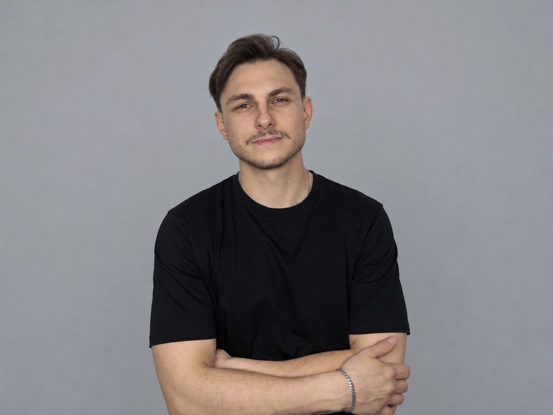 Дмитрий Меланьин в чёрной футболке со скрещёнными руками у серой стены