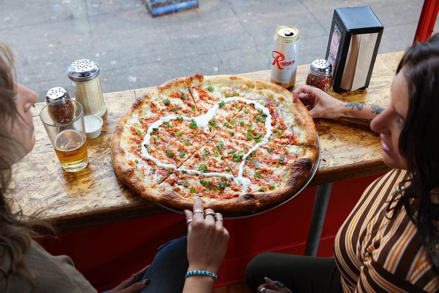 Two people sitting around a large pizza, with a drizzle of sauce in the shape of a heart, while they are drinking beers