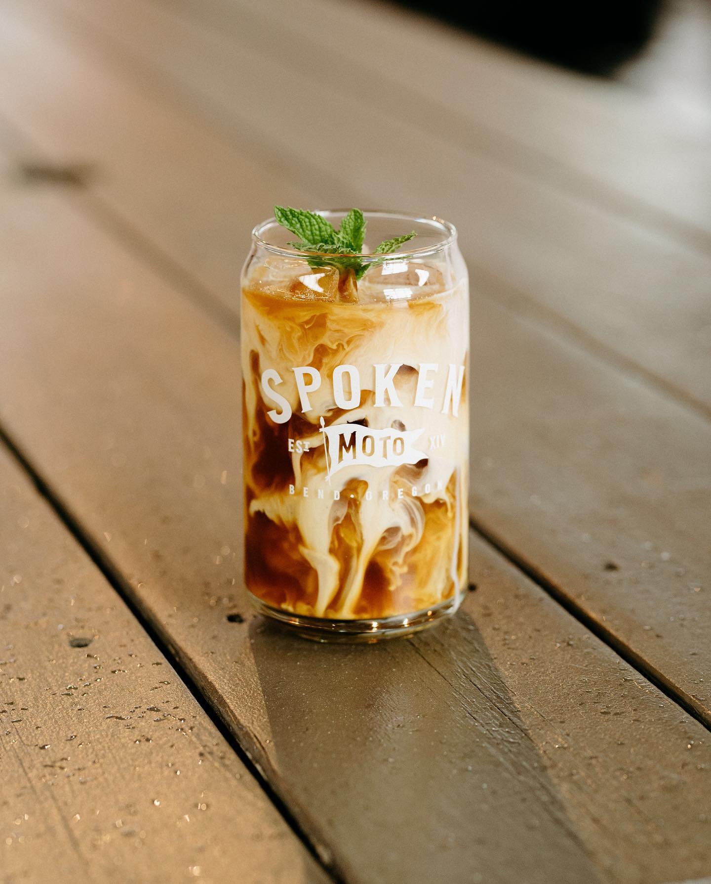 An beautiful iced coffee mixing with cream in a clear pint glass with a Spoken Moto logo on it. 