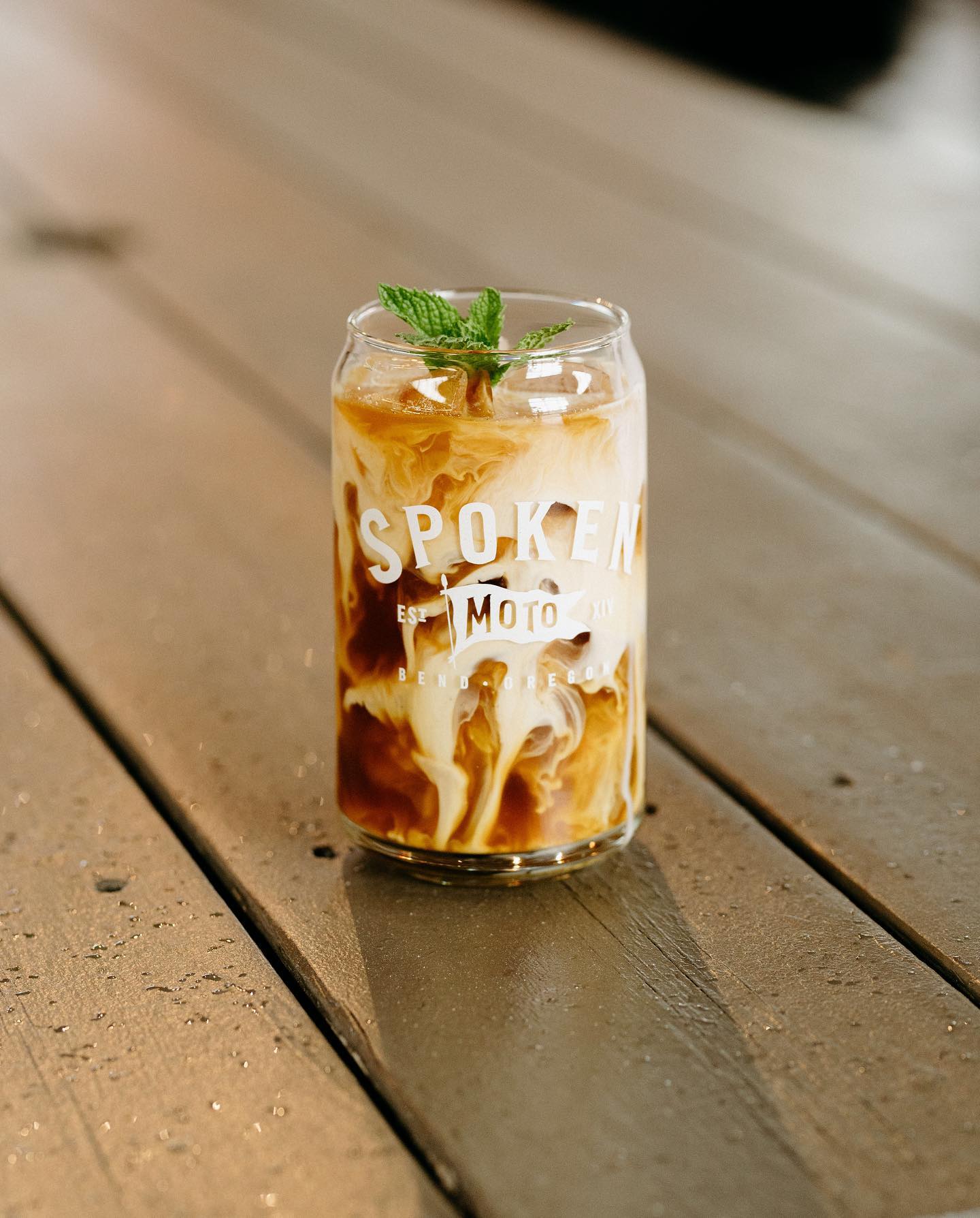 An beautiful iced coffee mixing with cream in a clear pint glass with a Spoken Moto logo on it.