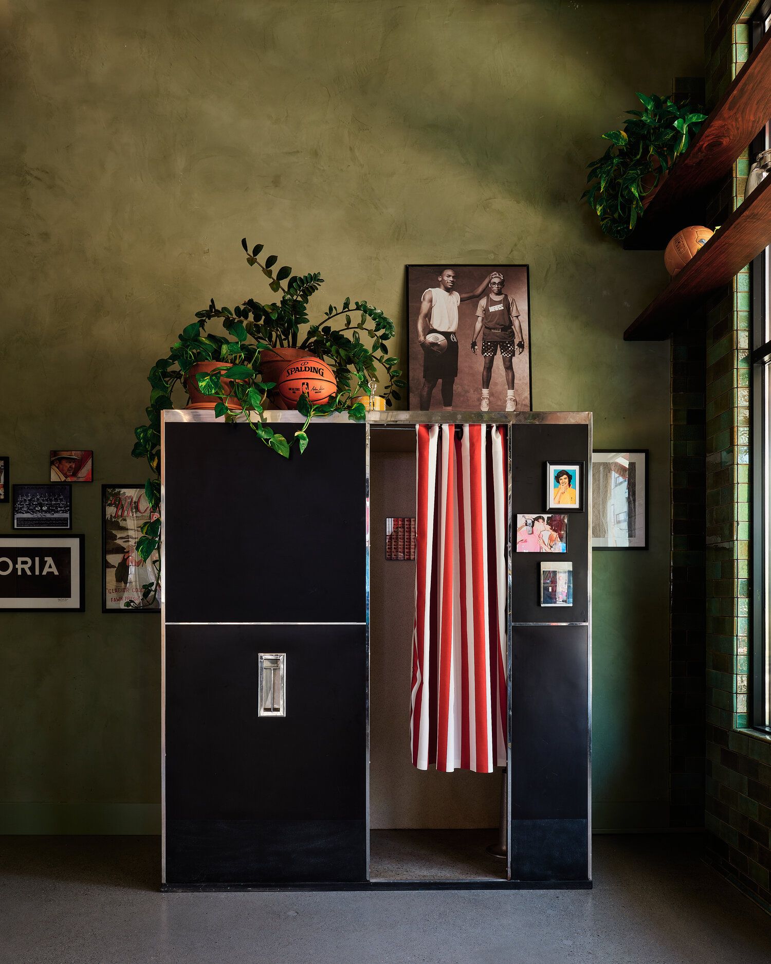 A vintage photobooth stands in front of a green limewashed wall in Cicoria. A portrait of basketball players and a plant sit atop the photobooth.