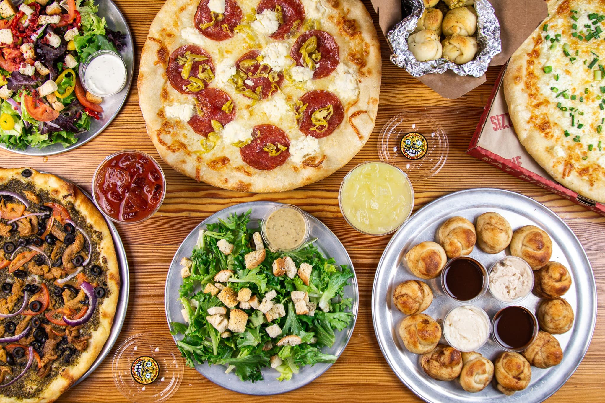 A top-down view of several pizzas, salads, garlic knots, and sauces
