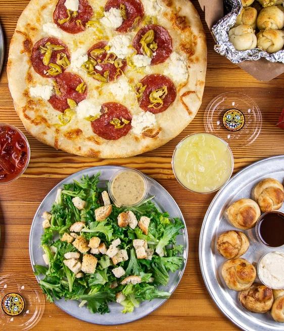 A top-down view of several pizzas, salads, garlic knots, and sauces