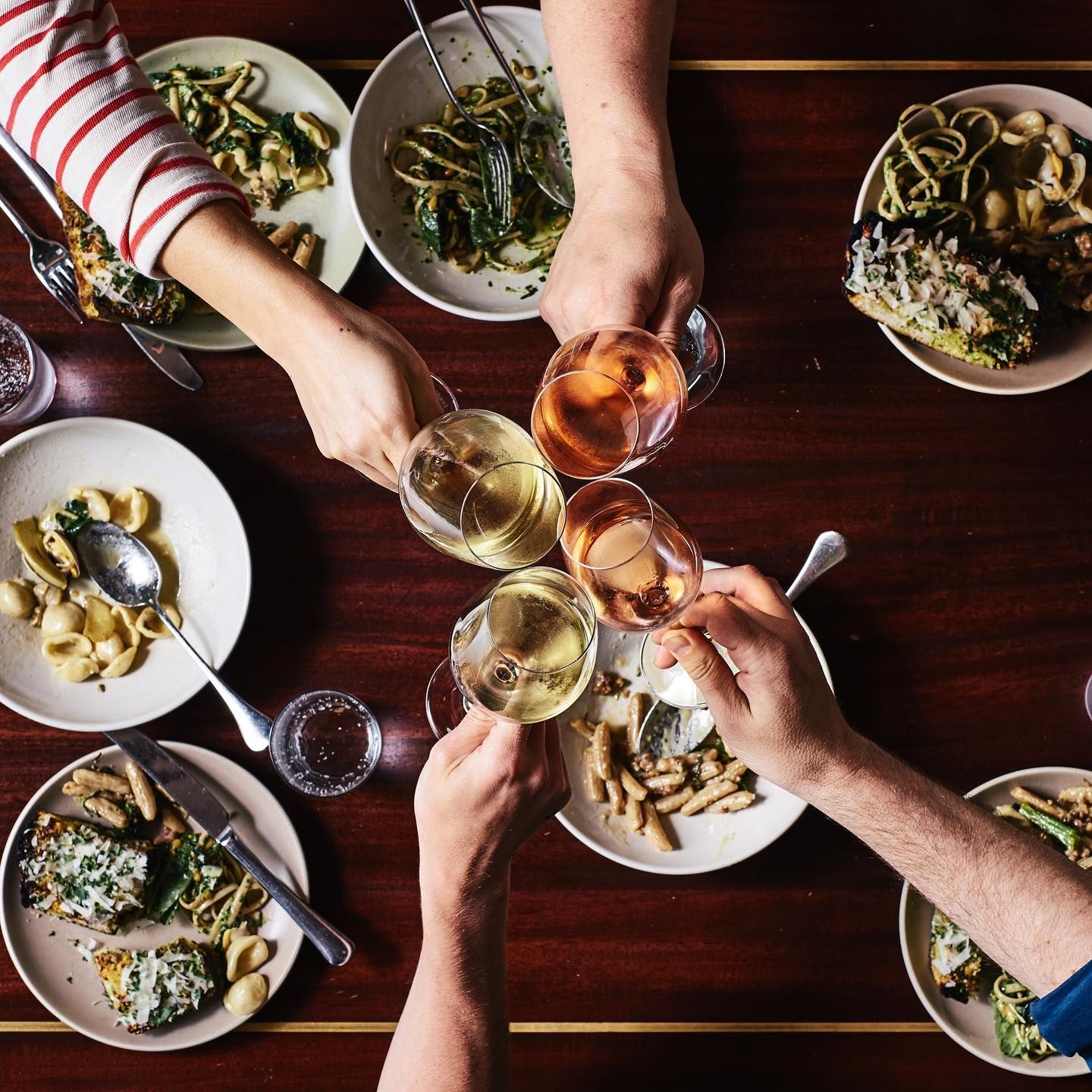A group of friends gather and cheers their wines over plates of food. 